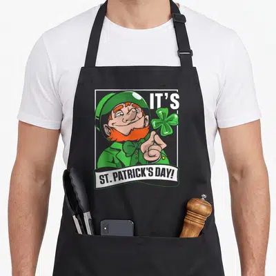 Tablier de cuisine noir avec illustration d'un lutin vert souriant tenant un trèfle à quatre feuilles et le texte It's St. Patrick's Day.