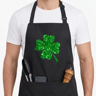 Tablier de cuisine noir avec un grand trèflic vert pailleté scintillant à quatre feuilles et une tige fine au centre.