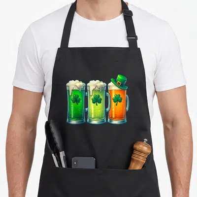 Tablier de cuisine noir avec trois verres de bière colorés, des trèfles verts et un petit chapeau de lutin sur le bord.