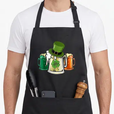 Tablier de cuisine noir avec illustration de trois pichets de bière et un chapeau vert surmonté d'un trèfle.