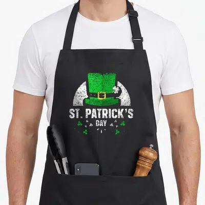 Tablier de cuisine noir avec un chapeau vert et l'inscription St Patrick's Day entourée de trèfles.