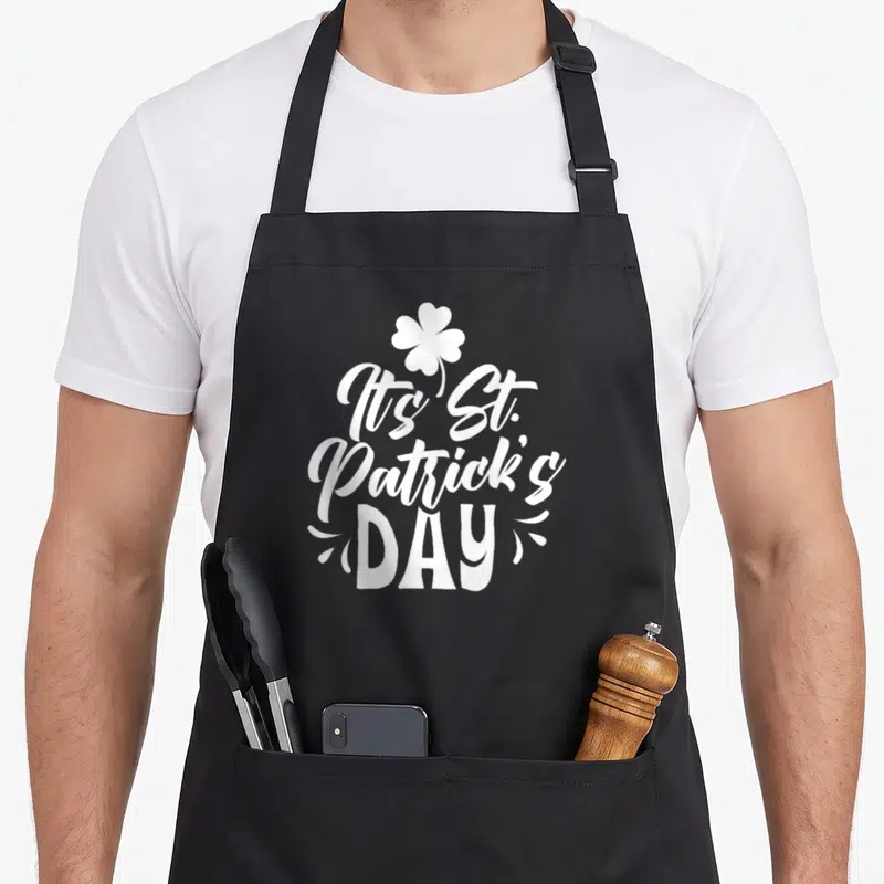 Tablier de cuisine noir avec le texte It's St. Patrick's Day et un trèfle blanc stylisé au centre.