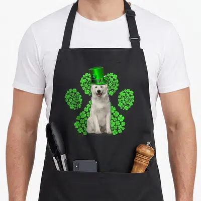 Tablier de cuisine noir avec un Berger Blanc Suisse en chapeau vert entouré de motifs de trèfles verts formant une patte.