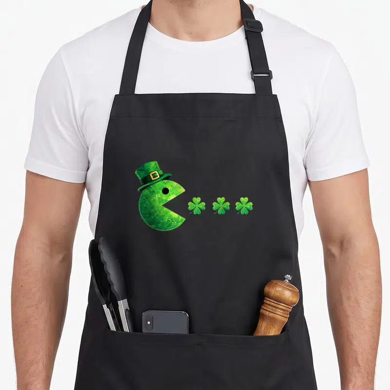 Tablier de cuisine noir avec pacman vert chapeau leprechaun dévorant trois trèfles à quatre feuilles alignés.