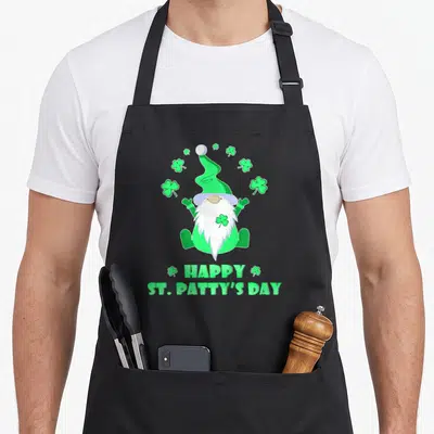 Tablier de cuisine noir avec un nain de jardin vert, des trèfles et le texte Happy St Patty's Day imprimé en bas.