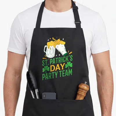 Tablier de cuisine noir avec deux verres de bière s'entrechoquant, le texte St. Patrick's Day Party Team et des trèfles verts.