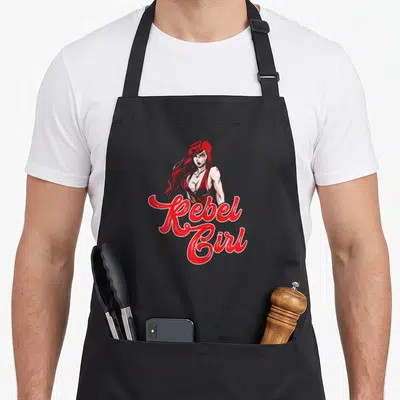 Tablier de cuisine noir avec illustration Rebel Girl rousse et texte rouge en calligraphie stylisée.