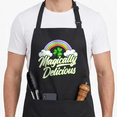Tablier de cuisine noir avec arc-en-ciel, trèfle à quatre feuilles et texte Magically Delicious en lettres blanches et vertes.