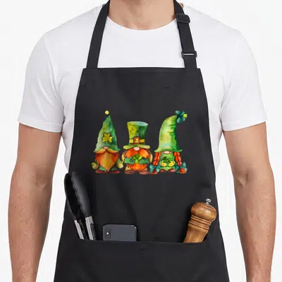 Tablier de cuisine noir avec trois gnomes aux chapeaux verts et des trèfles irlandais imprimés en aquarelle.