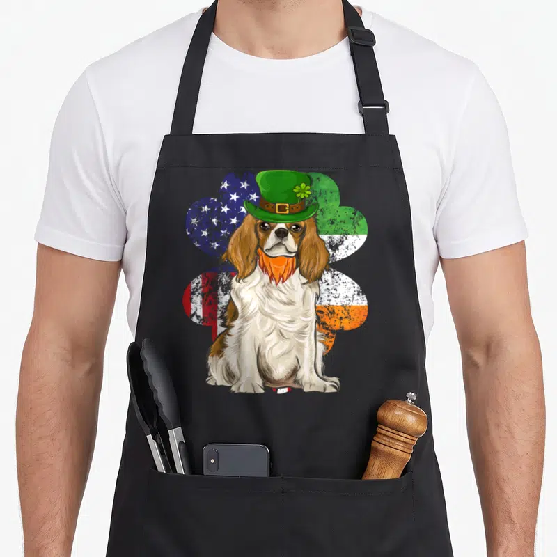 Tablier de cuisine noir avec illustration d'un chien Cavalier King Charles coiffé d'un chapeau vert sur fond de trèfle bicolore.