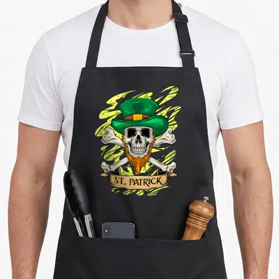 Tablier de cuisine noir avec un crâne irlandais au chapeau vert, barbe orange et os croisés sur fond de trèfles.