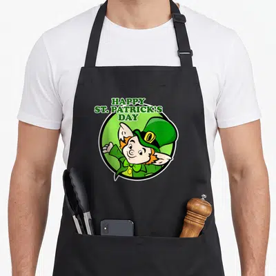 Tablier de cuisine noir avec un lutin aux cheveux roux et un chapeau vert, entouré du texte Happy St Patrick's Day.