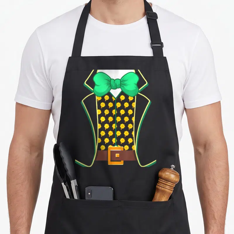 Tablier de cuisine noir avec illustration de costume de lutin, nœud papillon vert et motifs de trèfles jaunes sur le gilet.