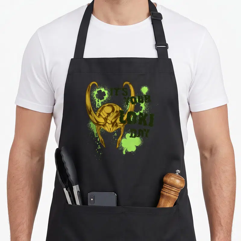 Tablier de cuisine noir avec casque doré de Loki et trèfles verts sur fond gris.