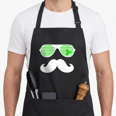Tablier de cuisine noir avec lunettes vertes rayées, trèfle et moustache blanche stylisée.