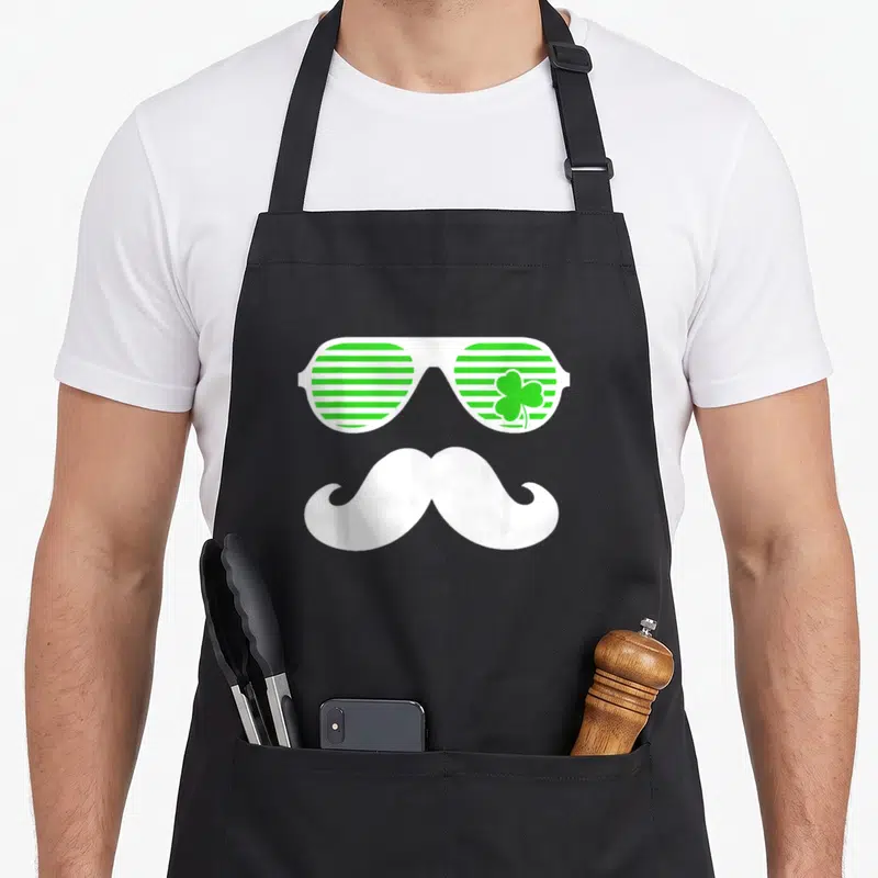 Tablier de cuisine noir avec lunettes vertes rayées, trèfle et moustache blanche stylisée.
