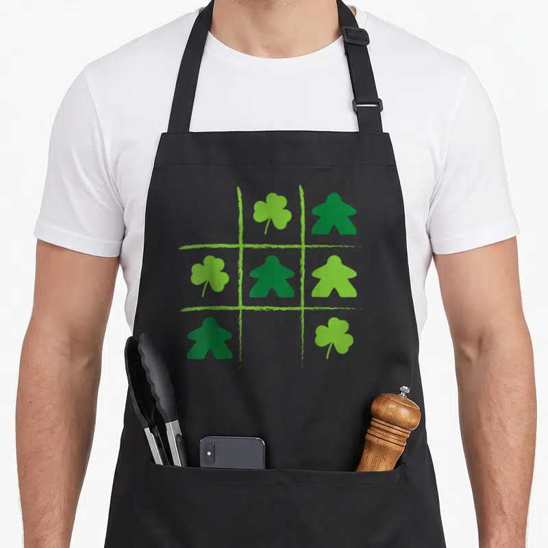 Tablier de cuisine noir avec une grille de tic-tac-toe mêlant des pions de jeu et des trèfles verts stylisés.