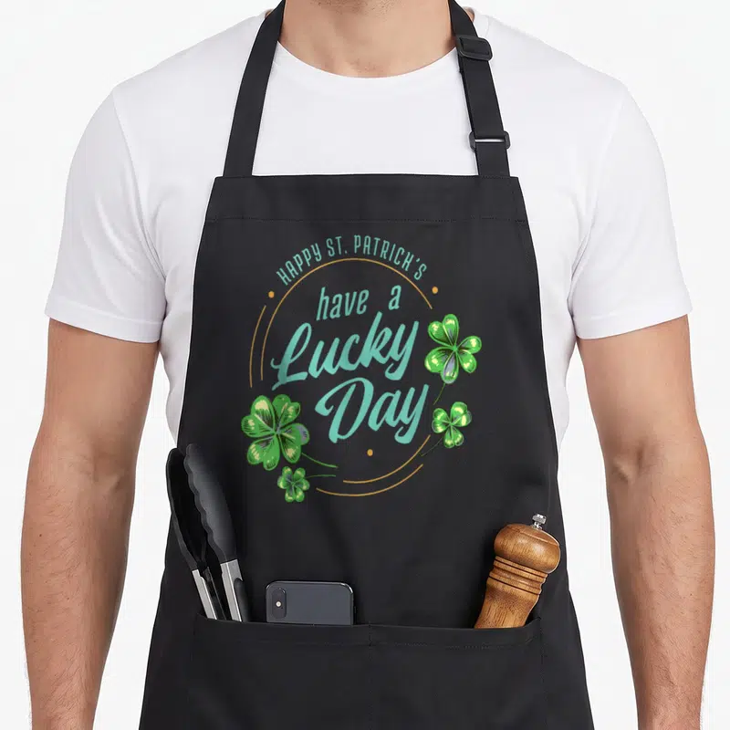 Tablier de cuisine noir avec inscription Happy St. Patrick's Have a Lucky Day et illustrations de trèfles verts.