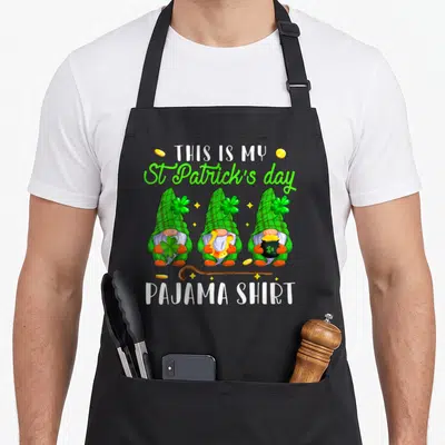 Tablier de cuisine noir avec trois gnomes verts, des trèfles, des pièces d'or et le texte This Is My St Patrick's Day Pajama Shirt imprimé en blanc et vert.