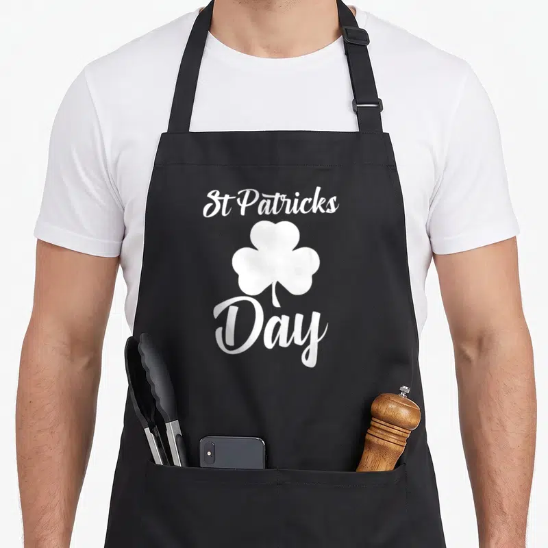 Tablier de cuisine noir avec inscription St Patricks Day et illustration de trèfle blanc au centre.