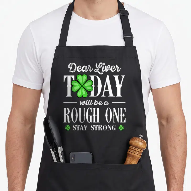 Tablier de cuisine noir avec le texte Dear Liver Today will be a rough one et des trèfles verts imprimés sur le devant.