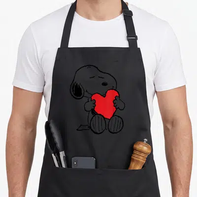 Tablier de cuisine noir avec Snopy assis tenant un grand cœur rouge vif dans ses pattes.