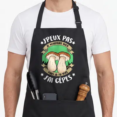 Tablier de cuisine noir avec illustration de deux cèpes et texte humoristique sur fond vert.