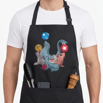 Tablier de cuisine noir avec illustration de quatre otaries et phoques jouant avec des balles colorées ornées d'étoiles blanches.