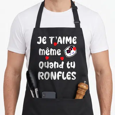 Tablier de cuisine noir avec texte Je t'aime même quand tu ronfles et illustration d'un visage dormant avec des cœurs rouges.
