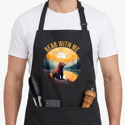Tablier de cuisine noir avec illustration circulaire d'un ours brun devant un lac et des montagnes au coucher de soleil.