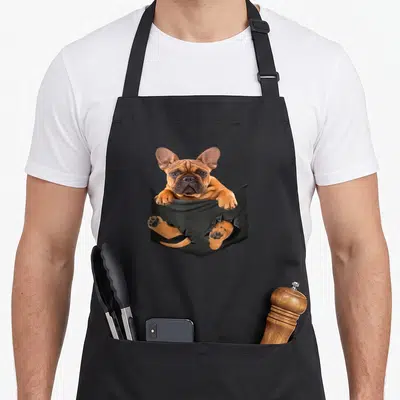 Tablier de cuisine noir avec illustration d'un bouledogue français marron émergeant d'une poche noire avec ses pattes et sa queue visibles.