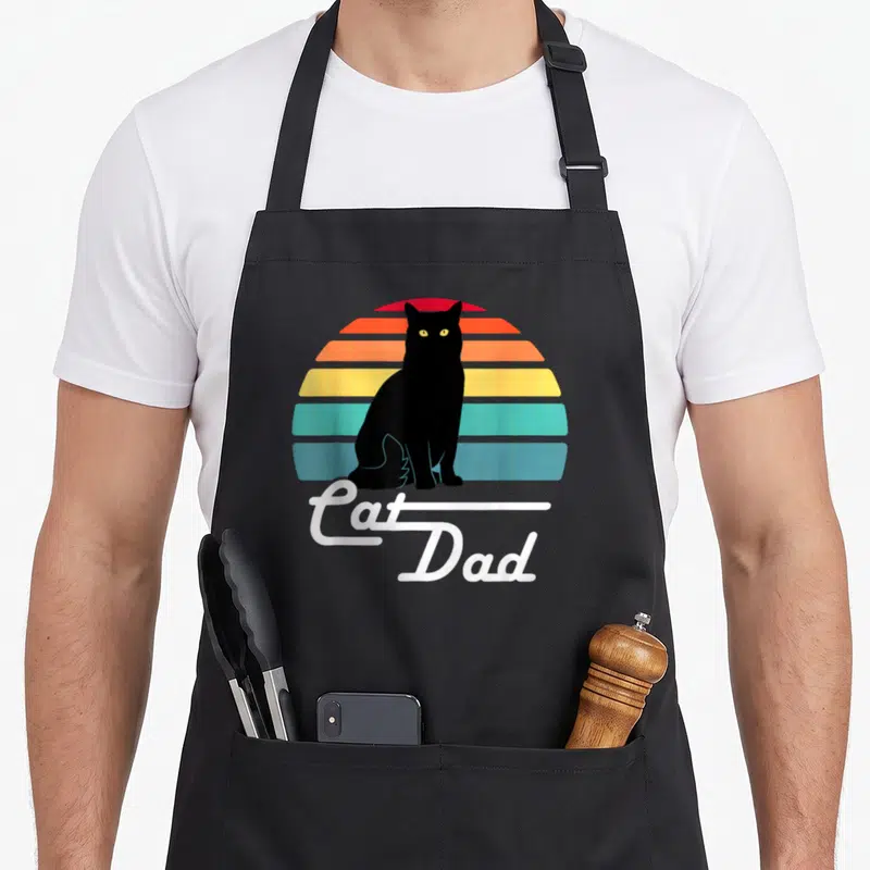 Tablier de cuisine noir avec un chat noir assis devant un soleil coloré et linscription Cat Dad en blanc.