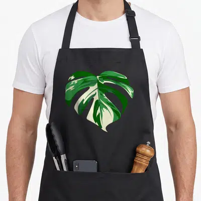 Tablier de cuisine noir avec une illustration colorée d'une feuille de Monstera Deliciosa Variegatas aux nuances de vert et de crème.