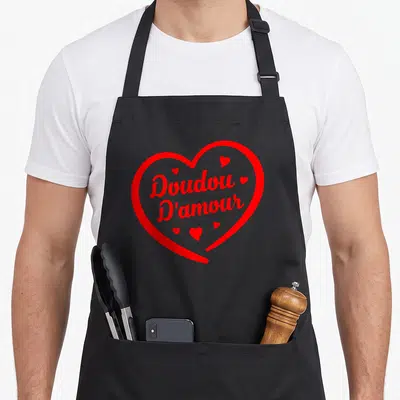 Tablier de cuisine noir orné d'un grand cœur rouge contenant le texte Doudou D'amour entouré de petits cœurs.