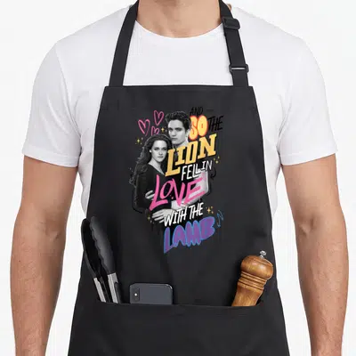 Tablier de cuisine noir avec illustration du couple et texte Le lion est tombé amoureux de l'agneau entouré de cœurs roses.