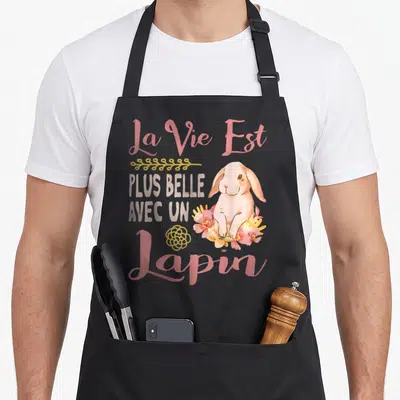 Tablier de cuisine noir avec un lapin rose entouré de fleurs et le texte La Vie Est Plus Belle Avec Un Lapin en lettres roses et grises.