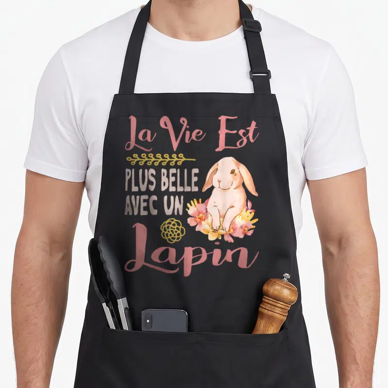 Tablier de cuisine noir avec un lapin rose entouré de fleurs et le texte La Vie Est Plus Belle Avec Un Lapin en lettres roses et grises.