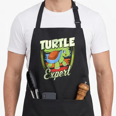 Tablier de cuisine noir avec une tortue verte souriante et le texte Turtle Expert en lettres capitales colorées.