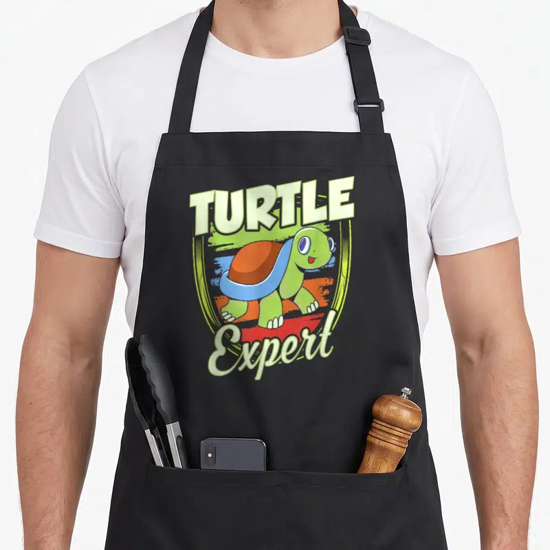 Tablier de cuisine noir avec une tortue verte souriante et le texte Turtle Expert en lettres capitales colorées.