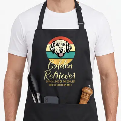 Tablier de cuisine noir avec tête de chien Golden Retriever sur fond circulaire coloré et texte doré.