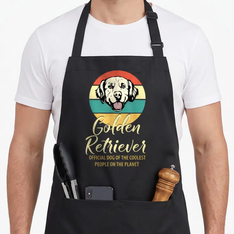Tablier de cuisine noir avec tête de chien Golden Retriever sur fond circulaire coloré et texte doré.