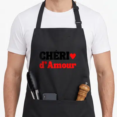 Tablier de cuisine noir avec l'inscription Chéri d'Amour en lettres noires et rouges accompagnée d'un petit cœur.