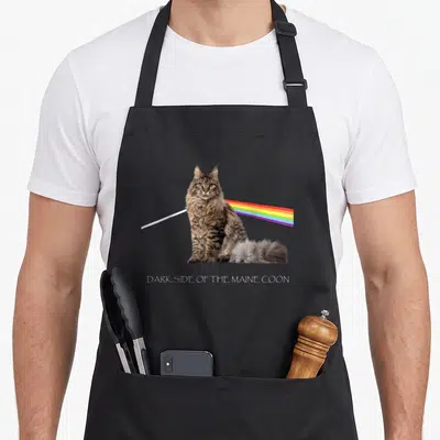 Tablier de cuisine noir avec un Maine Coon projetant un arc-en-ciel et le texte DARK SIDE OF THE MAINE COON en bas.