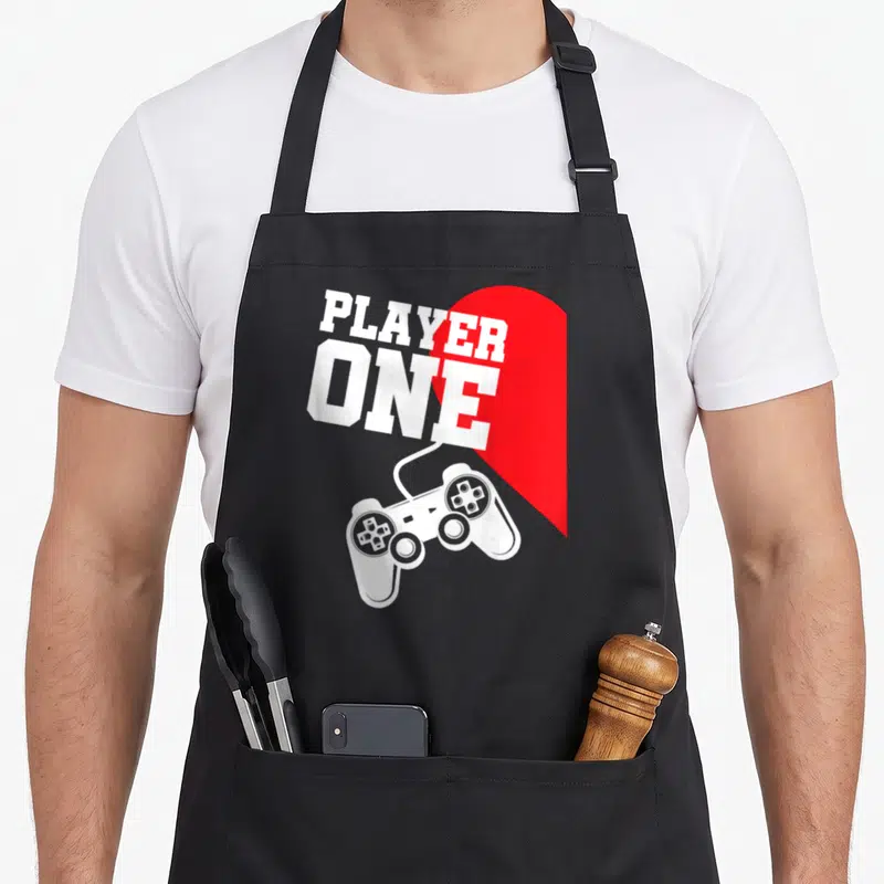 Tablier de cuisine noir avec inscription Player One, cœur rouge et manette de jeu vidéo blanche imprimée au centre.