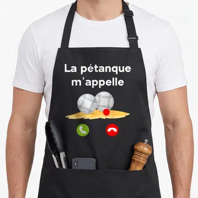 Tablier de cuisine noir avec l'inscription La pétanque m'appelle au-dessus de deux boules métalliques posées sur un tas de terre jaune.