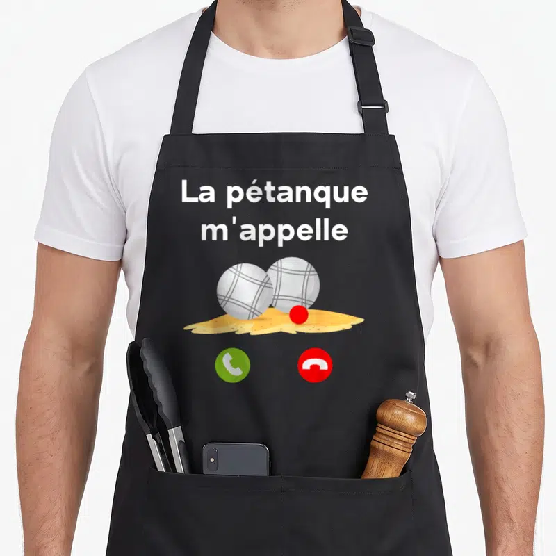 Tablier de cuisine noir avec l'inscription La pétanque m'appelle au-dessus de deux boules métalliques posées sur un tas de terre jaune.