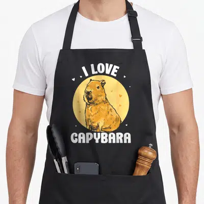 Tablier de cuisine noir avec illustration d'un capybara doré, cercle jaune, texte blanc I LOVE CAPYBARA et petits cœurs dispersés.
