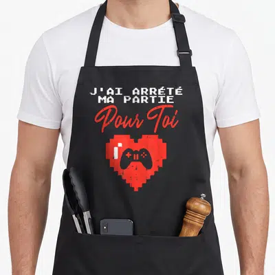 Tablier de cuisine noir avec texte blanc et rouge ainsi qu'un cœur pixelisé contenant une manette de jeu vidéo grise.