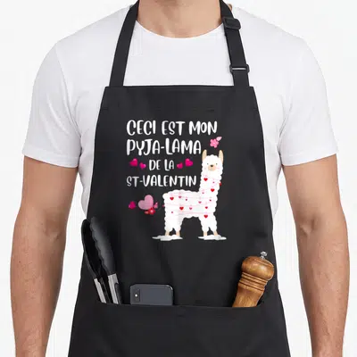 Tablier de cuisine noir avec illustration d'un lama blanc enroulé de ficelle rose et de petits cœurs, accompagné du texte ceci est mon pyja-lama de la st-valentin.