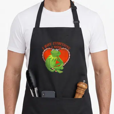 Tablier de cuisine noir avec Kermit le grenouille vert devant un cœur rouge et une inscription orange.